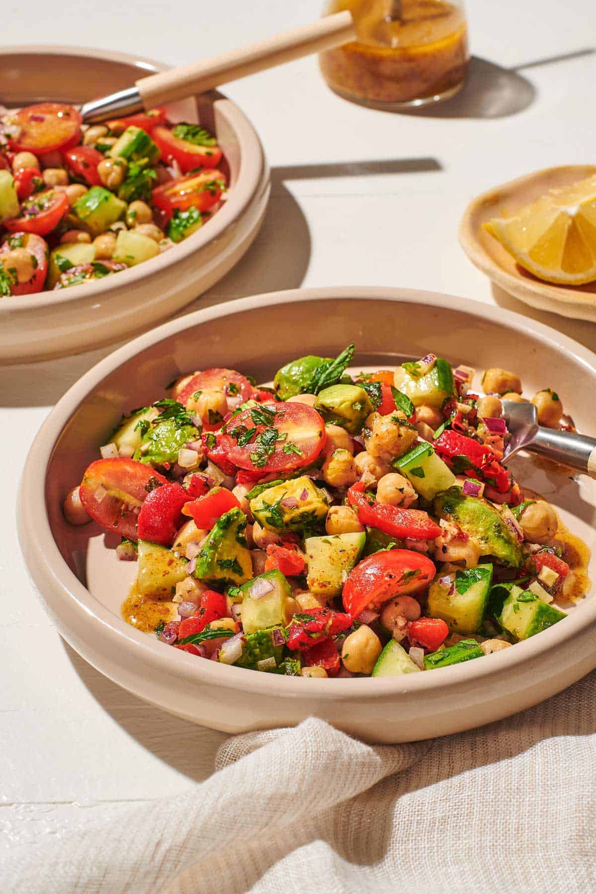 a close up of two bowls of vegan chickpea salad with forks next to a small bowl of lemon wedges, a jar of dijon dressing, and a cloth napkin.