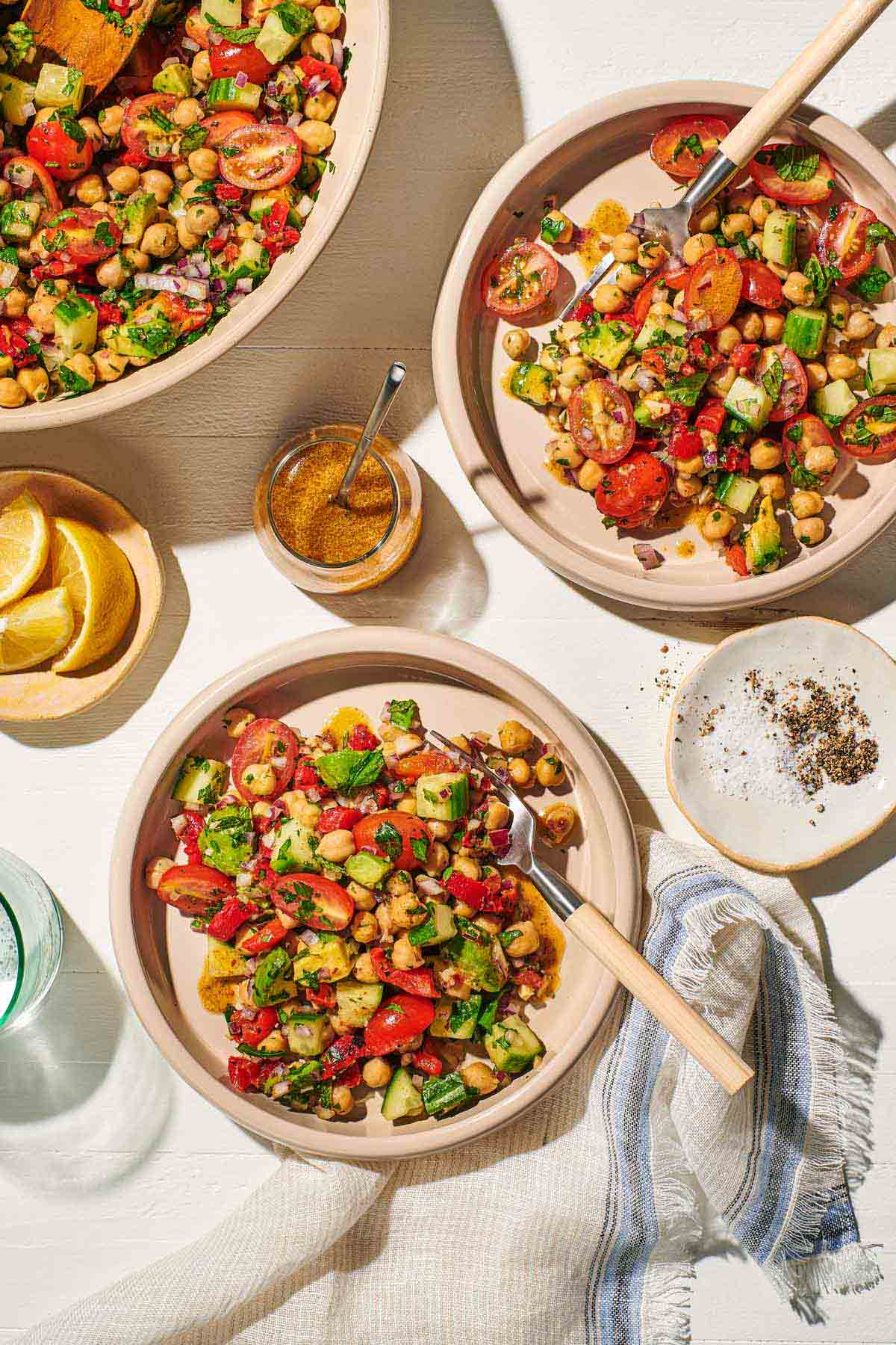 an overhead photo of two bowls of vegan chickpea salad with forks next to a serving bowl of the salad, a small plate of salt and pepper, a small bowl of lemon wedges, a jar of dijon dressing with a spoon, a glass of water and a cloth napkin.