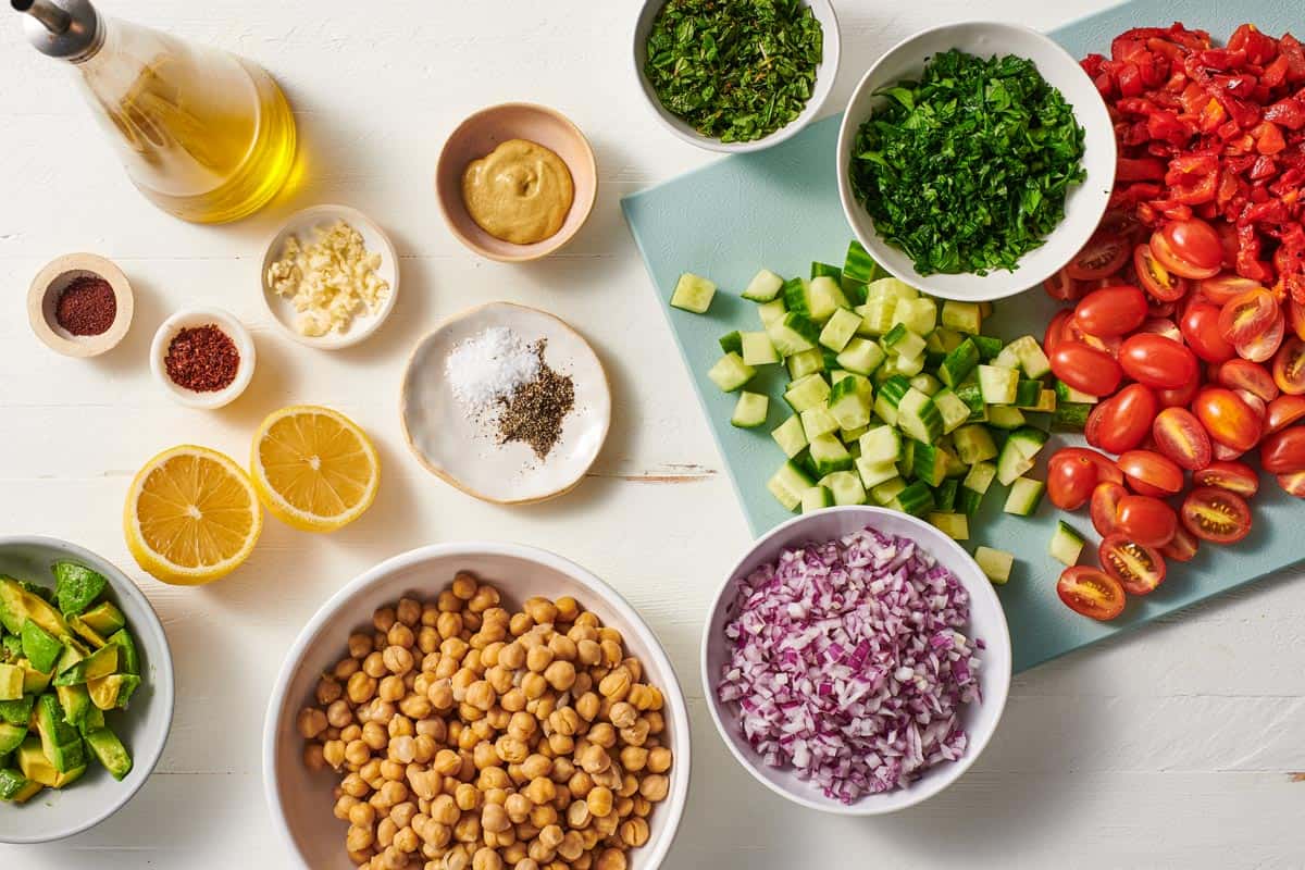 ingredients for vegan chickpea salad including dijon mustard, lemon, garlic, aleppo pepper, sumac, salt, pepper, olive oil, chickpeas, English cucumber, grape tomatoes, roasted red peppers, red onion, parsley, mint and avocado.