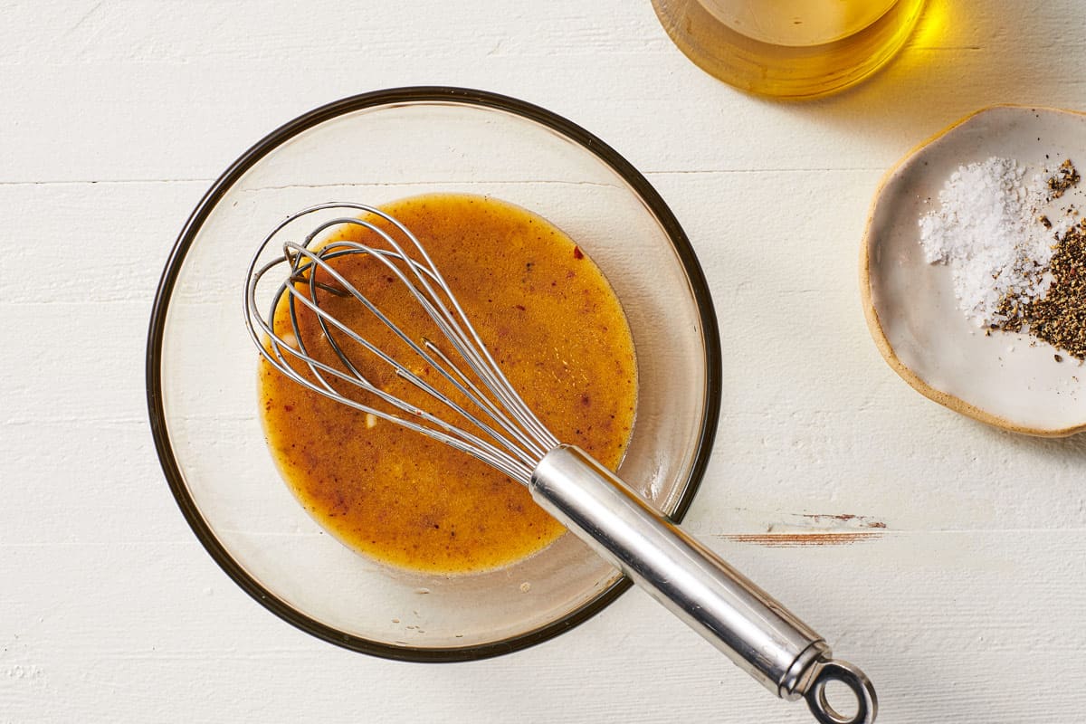 dijon dressing in a small mixing bowl with a whisk, next to a small plate of salt and pepper and a jar of olive oil.