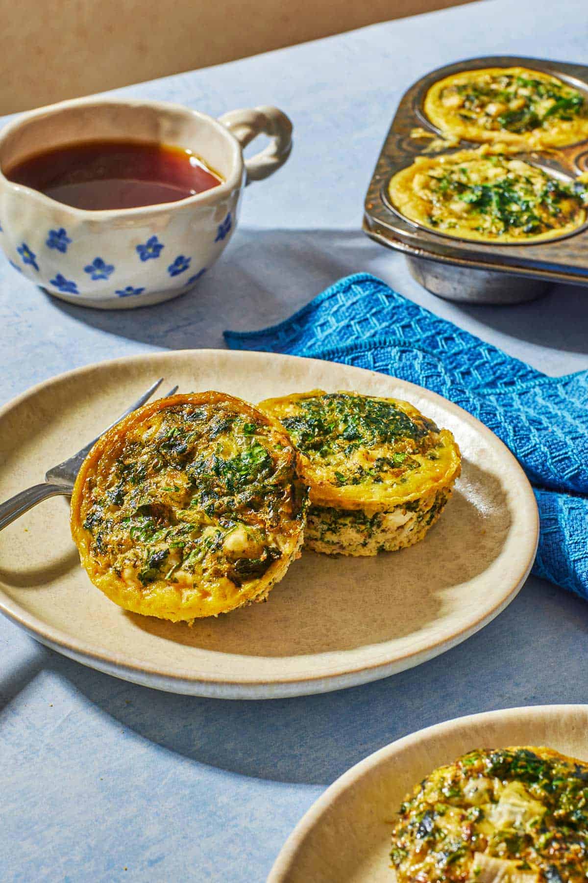 Two spanakopita egg muffins on a plate with a fork and a cup of tea in the background.