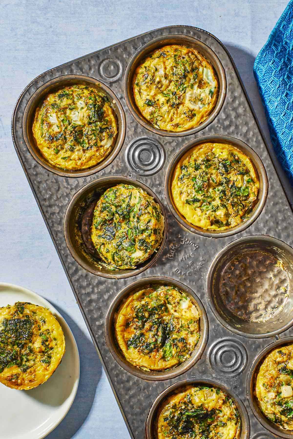 Overhead shot of Spanakopita egg muffins in a muffin tin, with one muffin on a plate alongside.