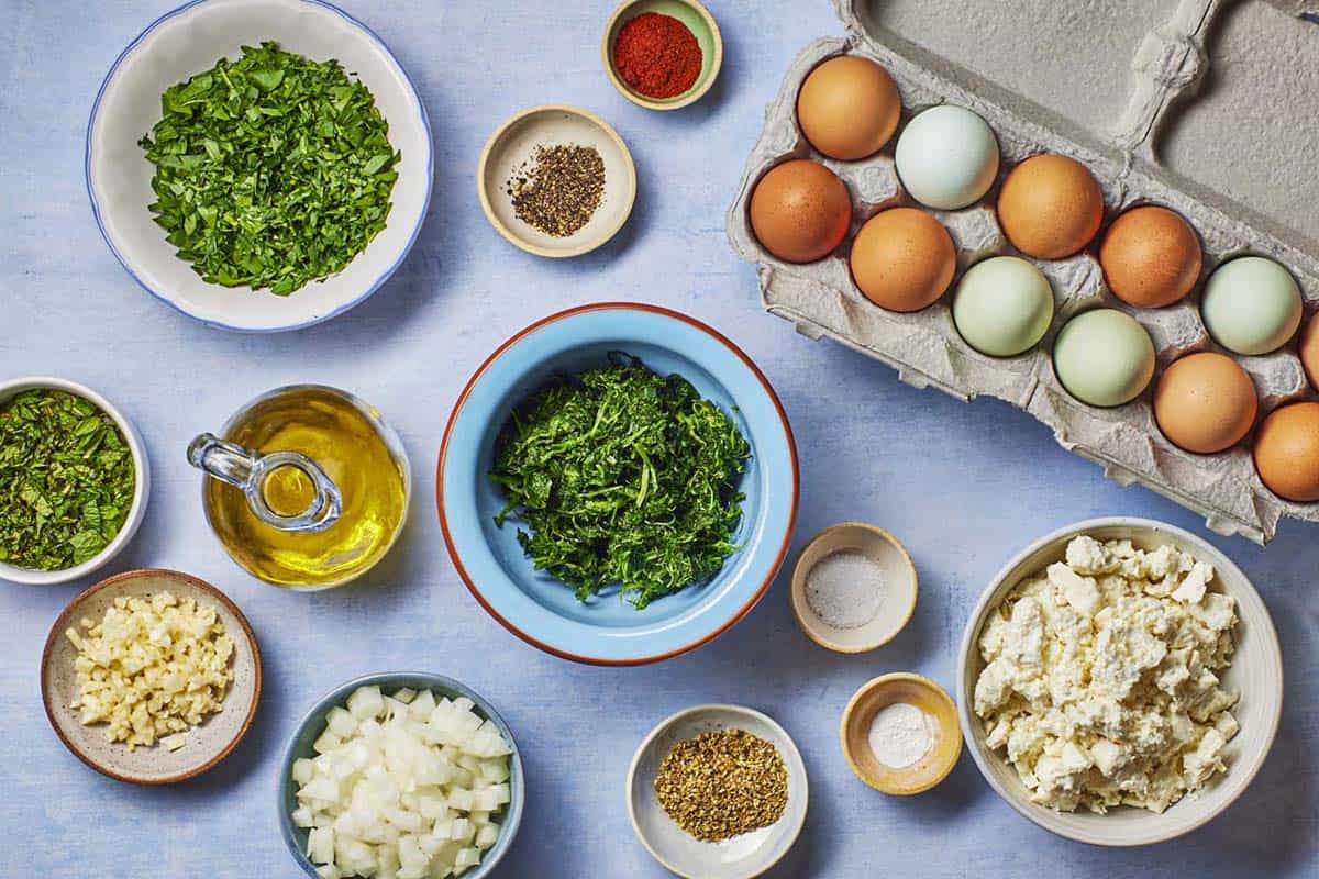 Ingredients for Spanakopita egg muffins, including olive oil, eggs, oregano, pepper, paprika, baking powder, salt, parsley, spinach, mint, garlic, and feta cheese.