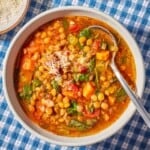 An overhead photo of a bowl of Italian lentil soup with a spoon next to a small bowl of grated parmesan cheese and olive oil.