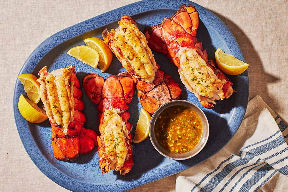 An overhead photo of 4 broiled lobster tails on a blue serving platter lemon wedges and butter sauce in a small bowl.