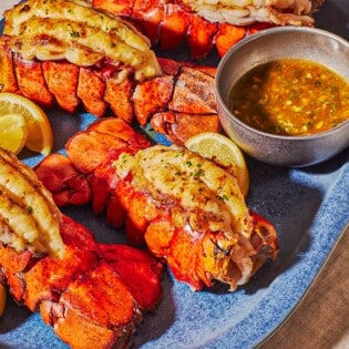 A close up of 4 broiled lobster tails on a blue serving platter lemon wedges and butter sauce in a small bowl.