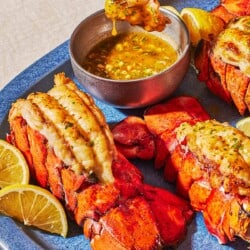 Three broiled lobster tails on a platter with lemon wedges and a bowl of butter sauce. A bite of lobster is being lifted from the bowl of butter sauce with a fork.