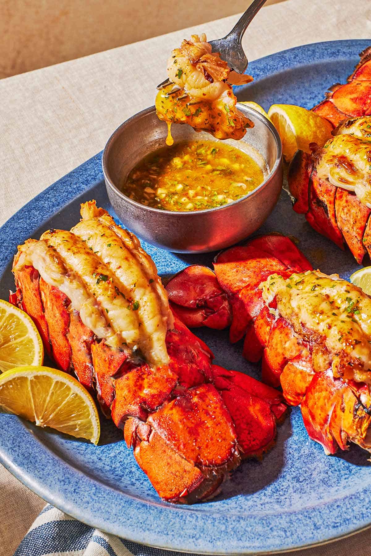 Three broiled lobster tails on a platter with lemon wedges and a bowl of butter sauce. A bite of lobster is being lifted from the bowl of butter sauce with a fork.