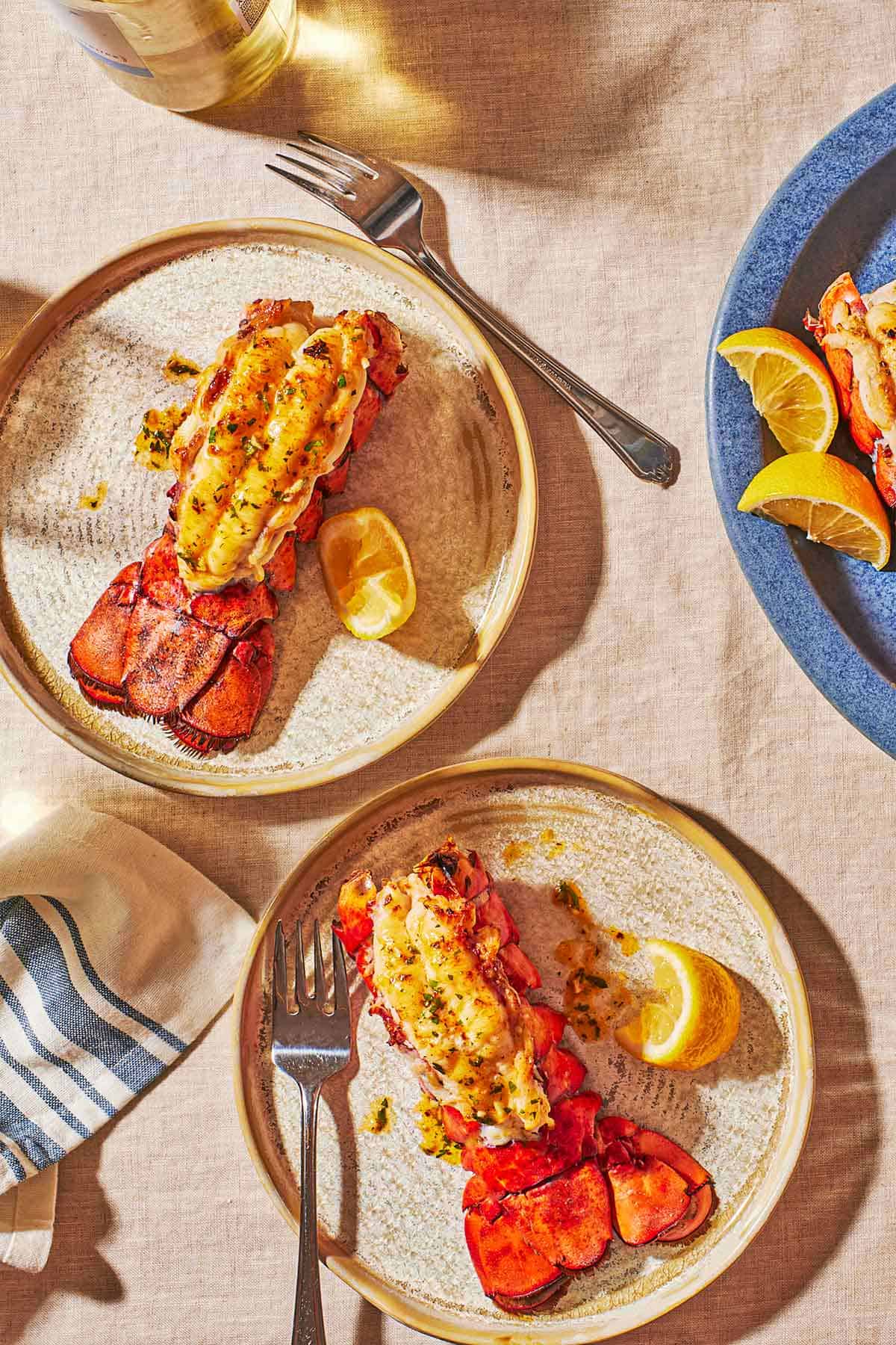 two plates with forks, each containing a broiled lobster tail and a lemon wedge.