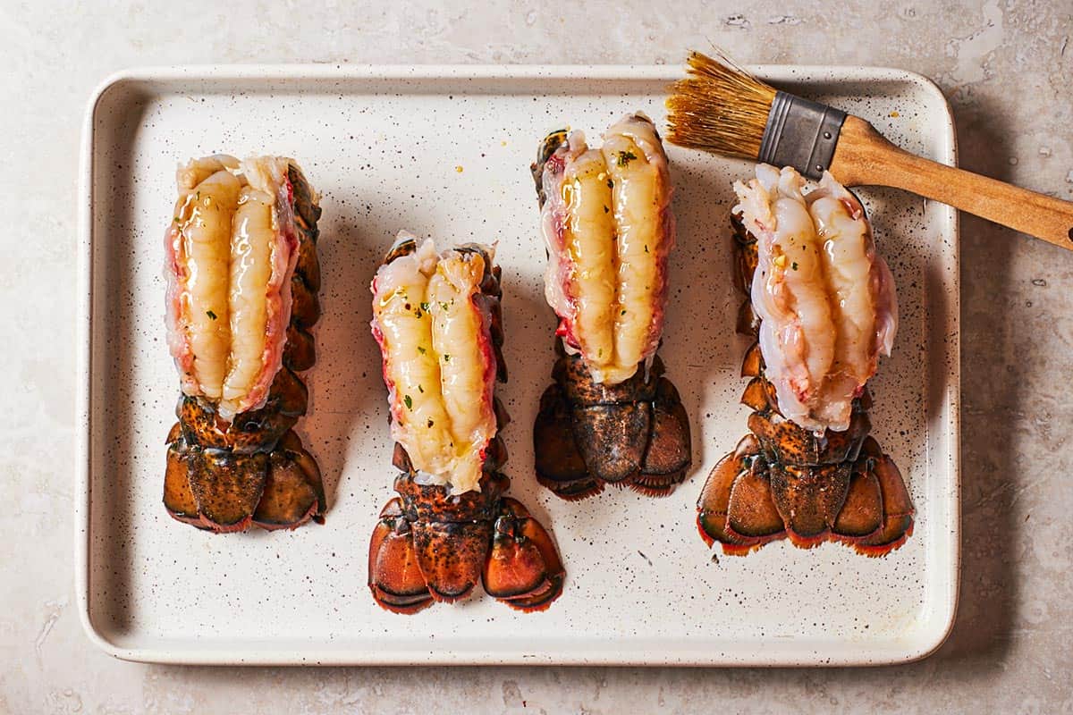 4 uncooked, butterflied lobster tails brushed with butter sauce on a sheet pan with a brush.