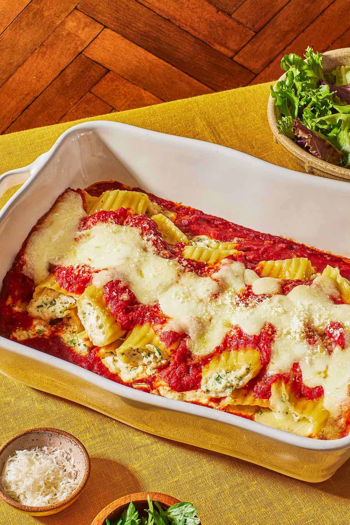 Several pieces of baked manicotti topped with spaghetti sauce and mozzarella in a baking dish next to a bowls of salad, grated parmesan cheese and parsley.