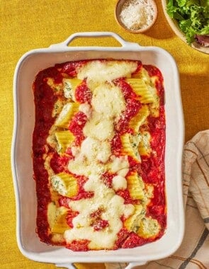 Several pieces of baked manicotti topped with spaghetti sauce and mozzarella in a baking dish next to a bowls of salad, and grated parmesan cheese.
