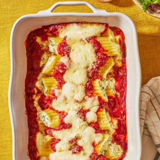 Several pieces of baked manicotti topped with spaghetti sauce and mozzarella in a baking dish next to a bowls of salad, and grated parmesan cheese.