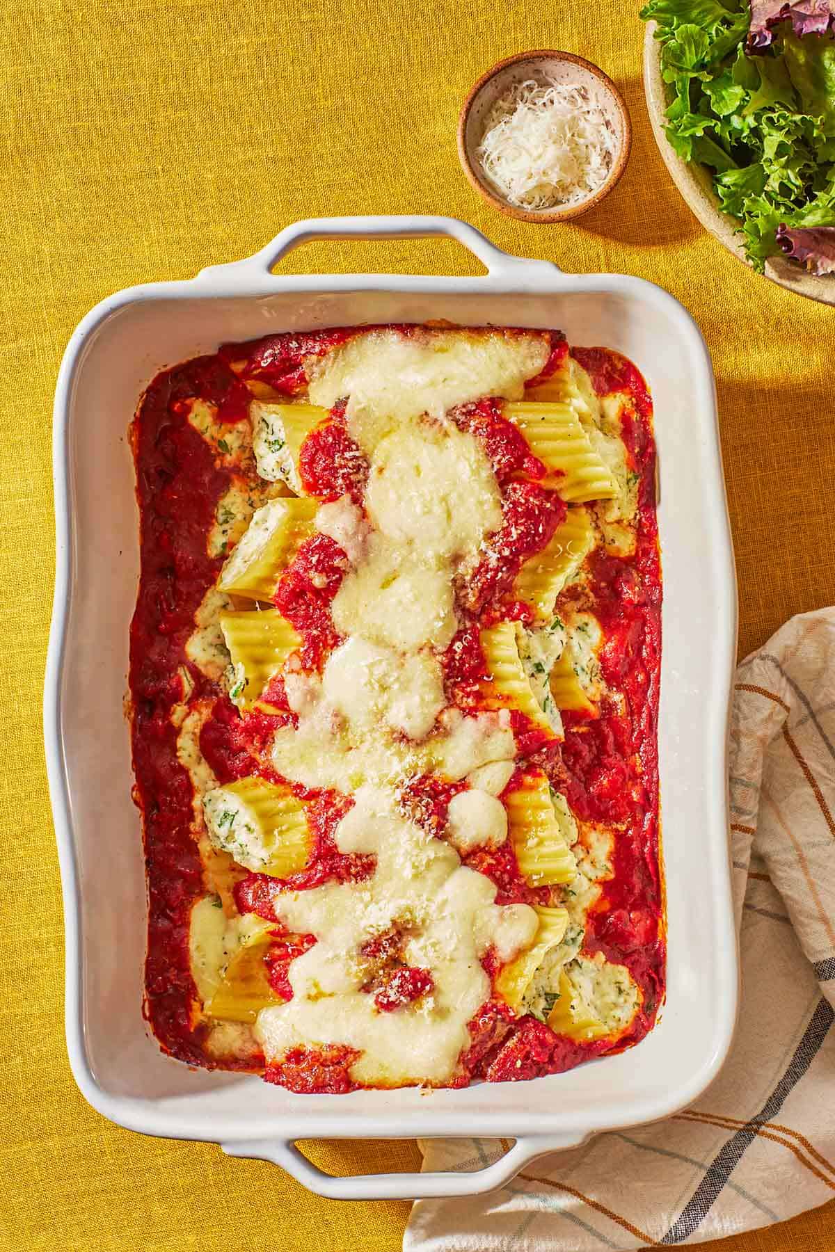Several pieces of baked manicotti topped with spaghetti sauce and mozzarella in a baking dish next to a bowls of salad, and grated parmesan cheese.