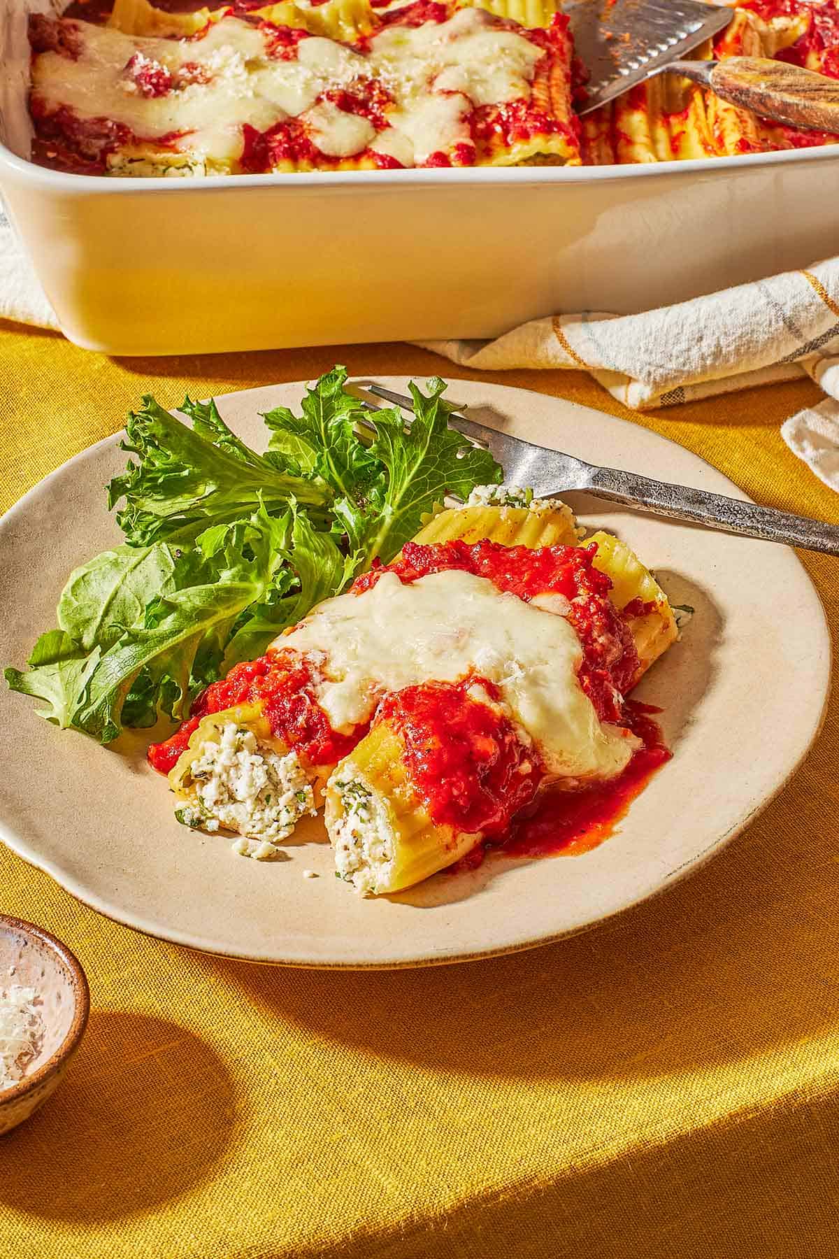 2 pieces of baked manicotti topped with spaghetti sauce and mozzarella cheese on a plate with a salad and a fork in front of a baking dish with the rest of the manicotti.
