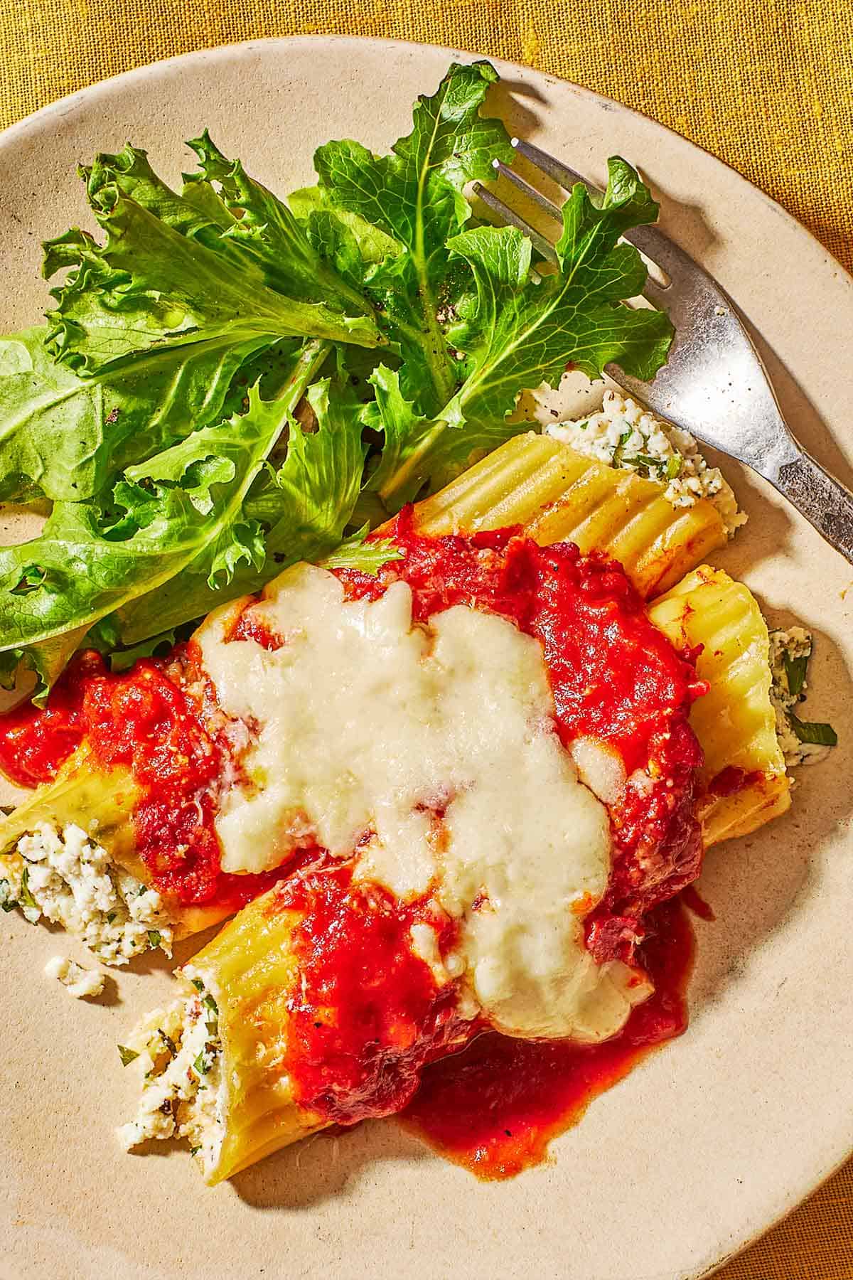 A close up of 2 pieces of baked manicotti topped with spaghetti sauce and mozzarella cheese on a plate with a salad and a fork.