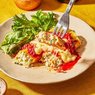 A fork picking up a bite of manicotti from a plate of more manicotti and a salad.