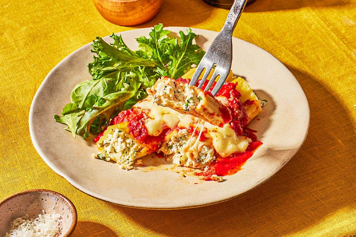 A fork picking up a bite of manicotti from a plate of more manicotti and a salad.