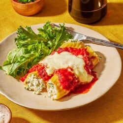 2 pieces of baked manicotti topped with spaghetti sauce and mozzarella cheese on a plate with a salad and a fork next to a glass of red wine and a small bowl of parsley.