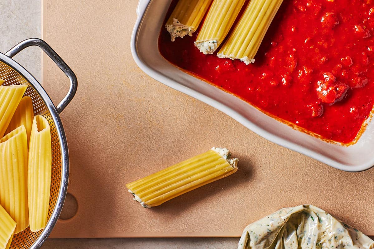 3 pieces of filled, unbaked manicotti on top of a layer of spaghetti sauce in a baking dish next to another filled manicotti and a colander of unfilled manicotti.