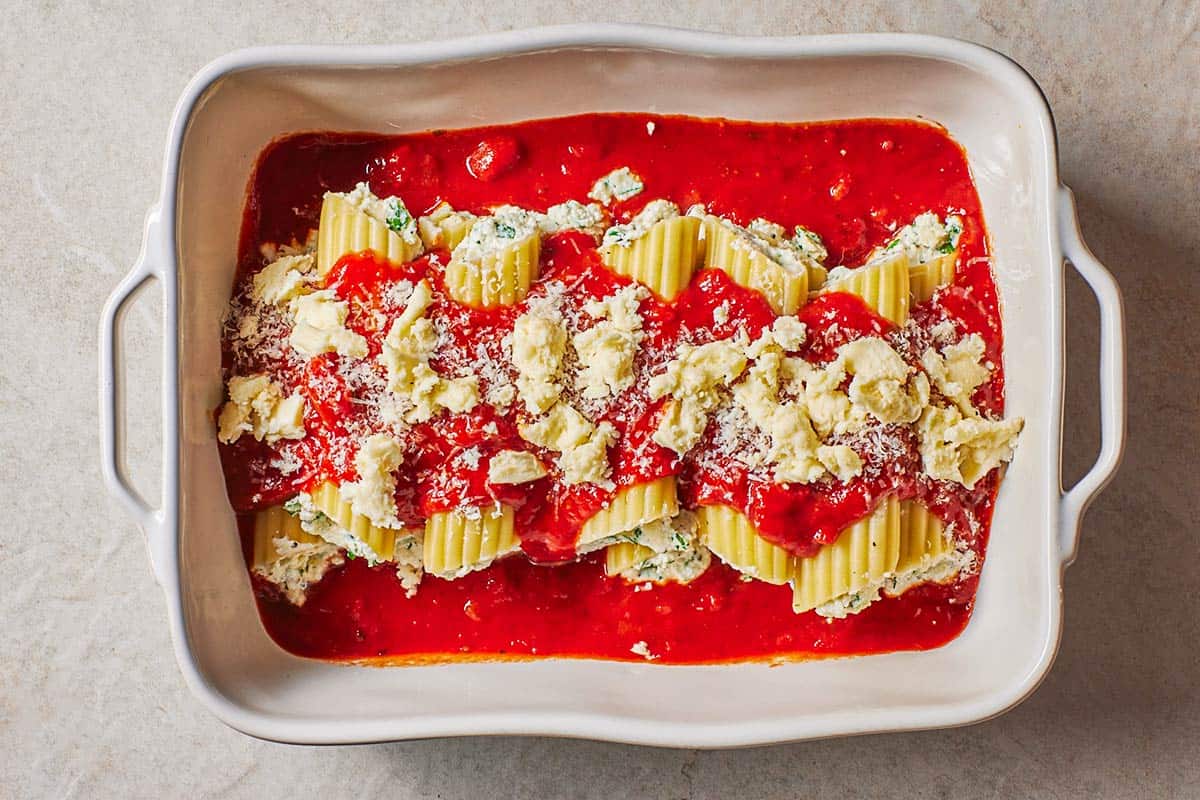 Several pieces of unbaked manicotti topped with spaghetti sauce and torn pieces of mozzarella cheese in a baking dish.