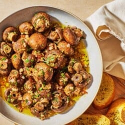 marinated mushrooms in a serving bowl next to several slices of crusty bread and two small picks.