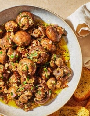 marinated mushrooms in a serving bowl next to several slices of crusty bread and two small picks.