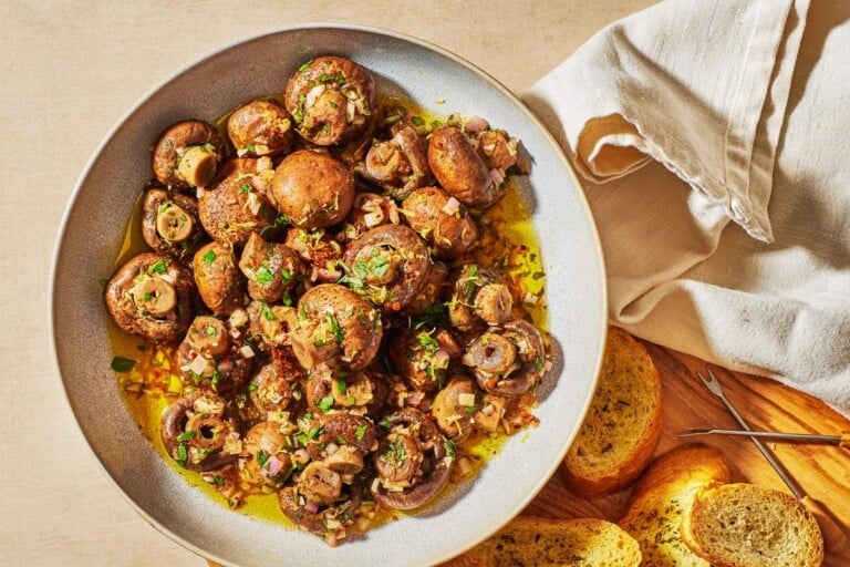 marinated mushrooms in a serving bowl next to several slices of crusty bread and two small picks.