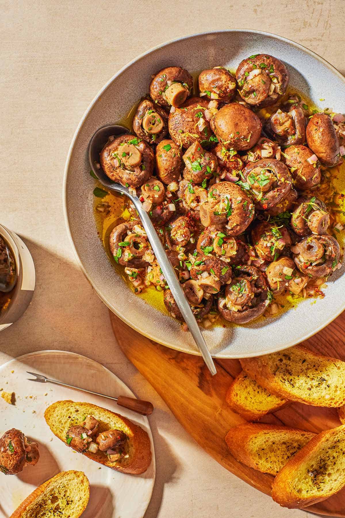 marinated mushrooms in a serving bowl with a spoon next to several slices of crusty bread and a plate with two slices of the bread and a small amount of marinated mushrooms.