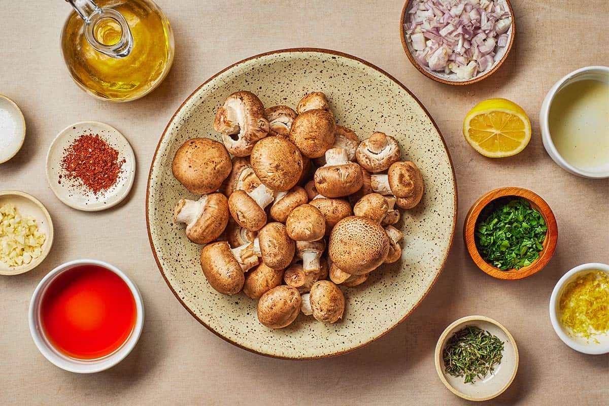 ingredients for marinated mushrooms including baby bella mushrooms, salt, minced shallots, parsley, thyme, aleppo pepper, lemon zest, lemon, red wine vinegar and olive oil.