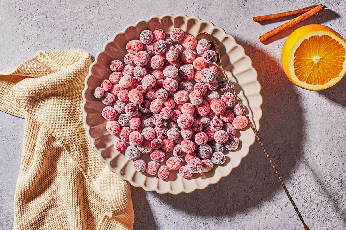 sugared cranberries in a serving bowl with a spoon next to a cloth napkin, an orange and 2 cinnamon sticks.