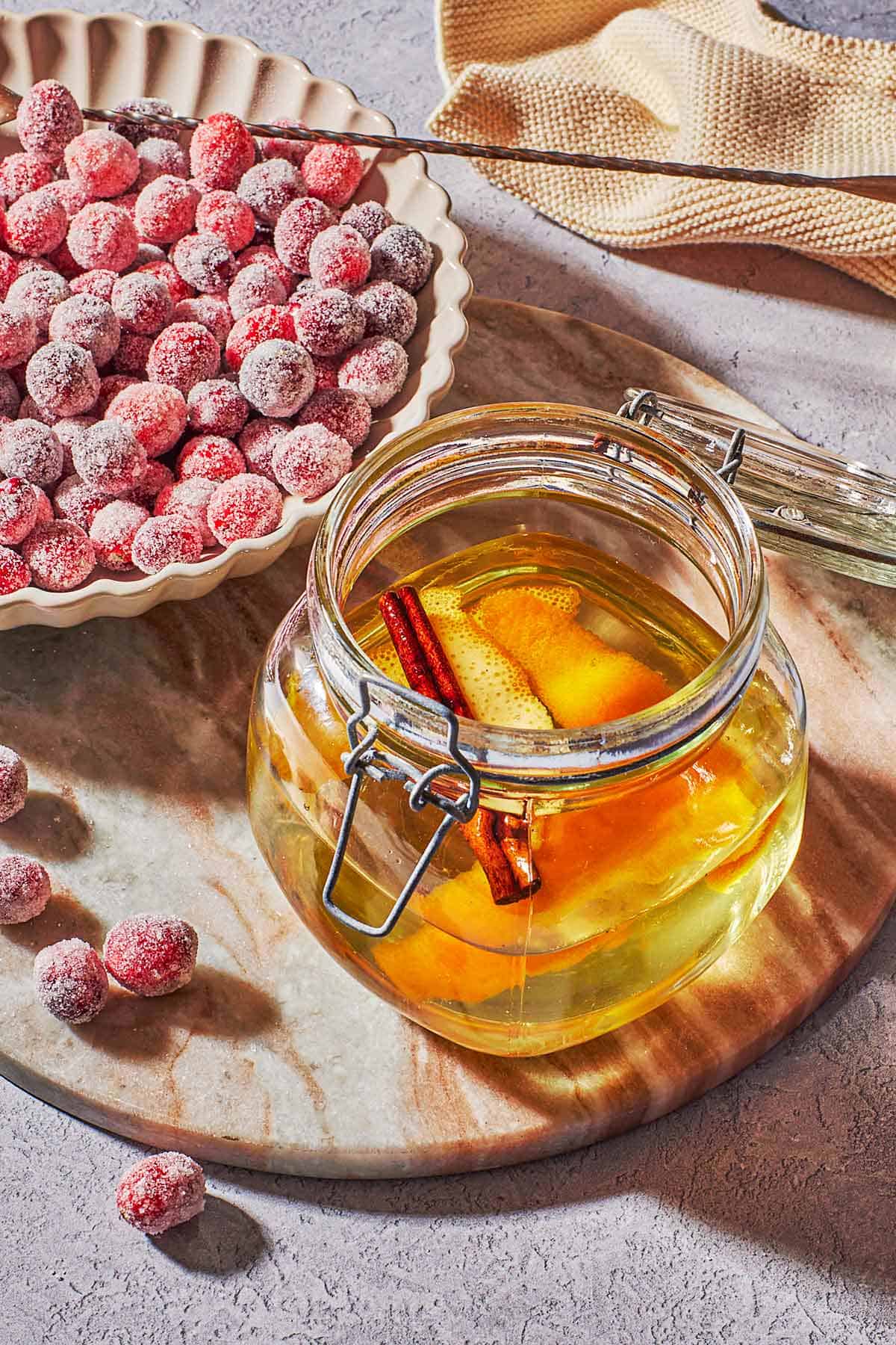 a bowl of sugared cranberries with a spoon next to a jar containing an orange peel and the cinnamon stick in simple syrup in a jar.