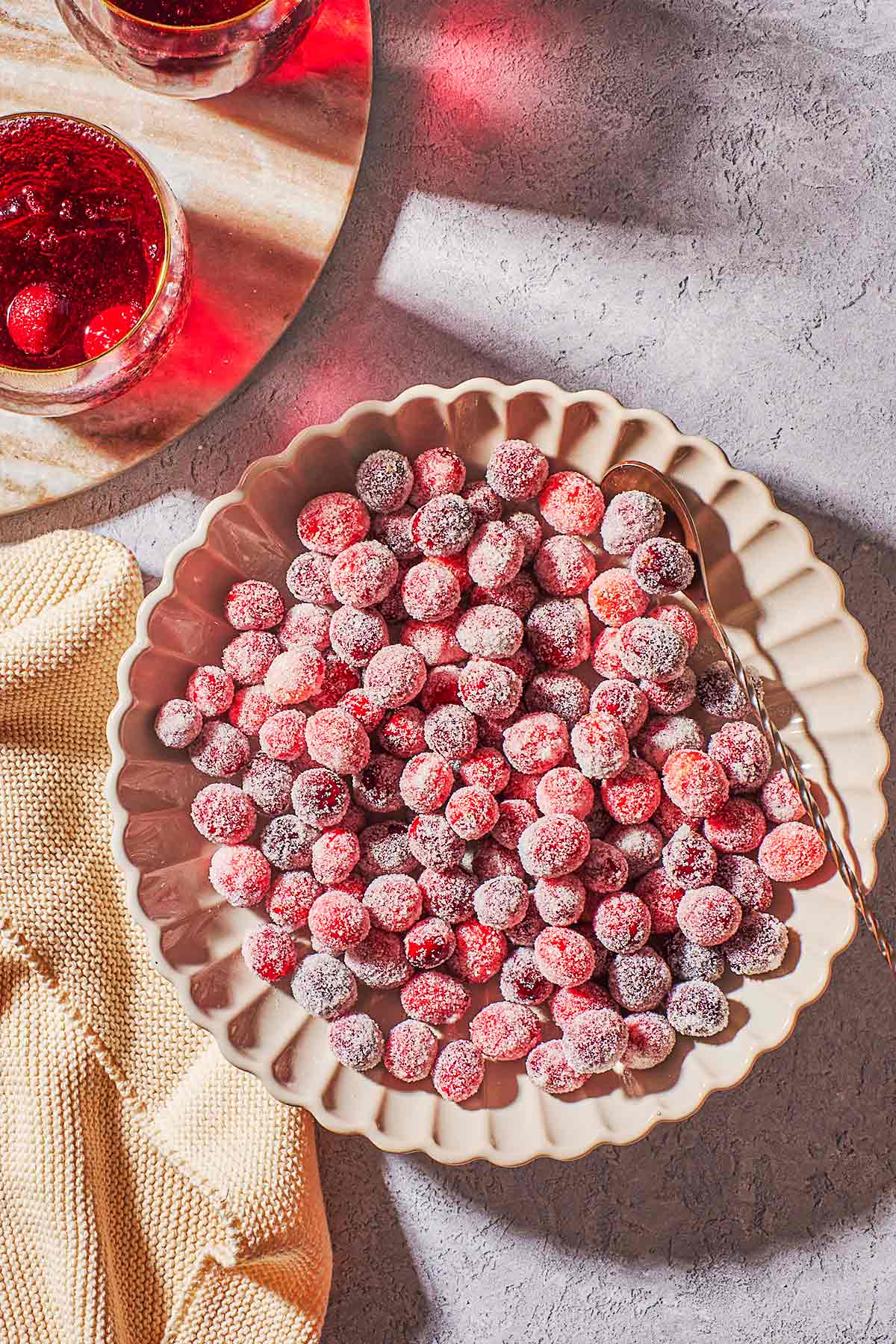 an overhead photo of sugared cranberries in a serving bowl with a spoon next to a cloth napkin and two drinks.