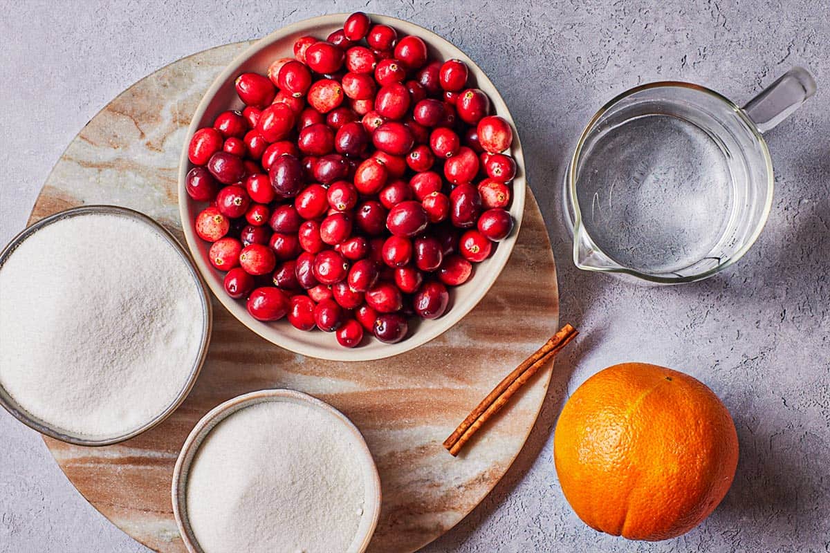 ingredients for sugared cranberries including fresh cranberries, an orange, granulated sugar, water, a cinnamon stick and superfine sugar.