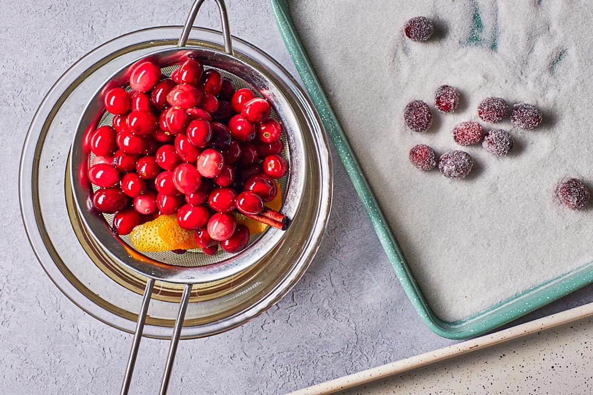 the cranberries, orange peel and cinnamon stick being drained in a mesh strainer placed over a bowl next to a baking sheet containing the superfine sugar and a few cranberries, already coated.