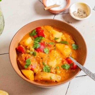 A bowl of vegetable stew with a spoon next to small bowls of salt and pepper.