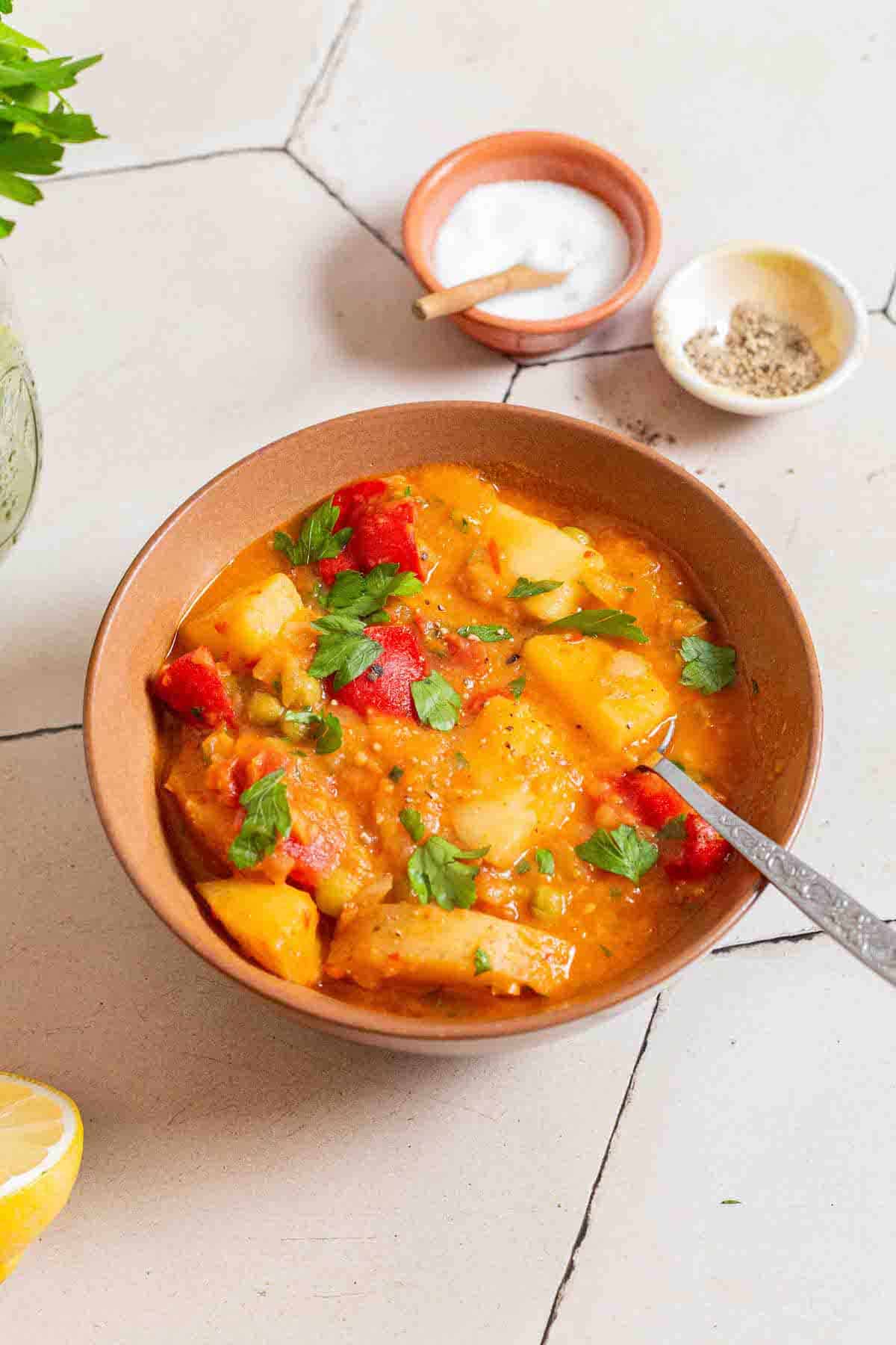 A bowl of vegetable stew with a spoon next to small bowls of salt and pepper.