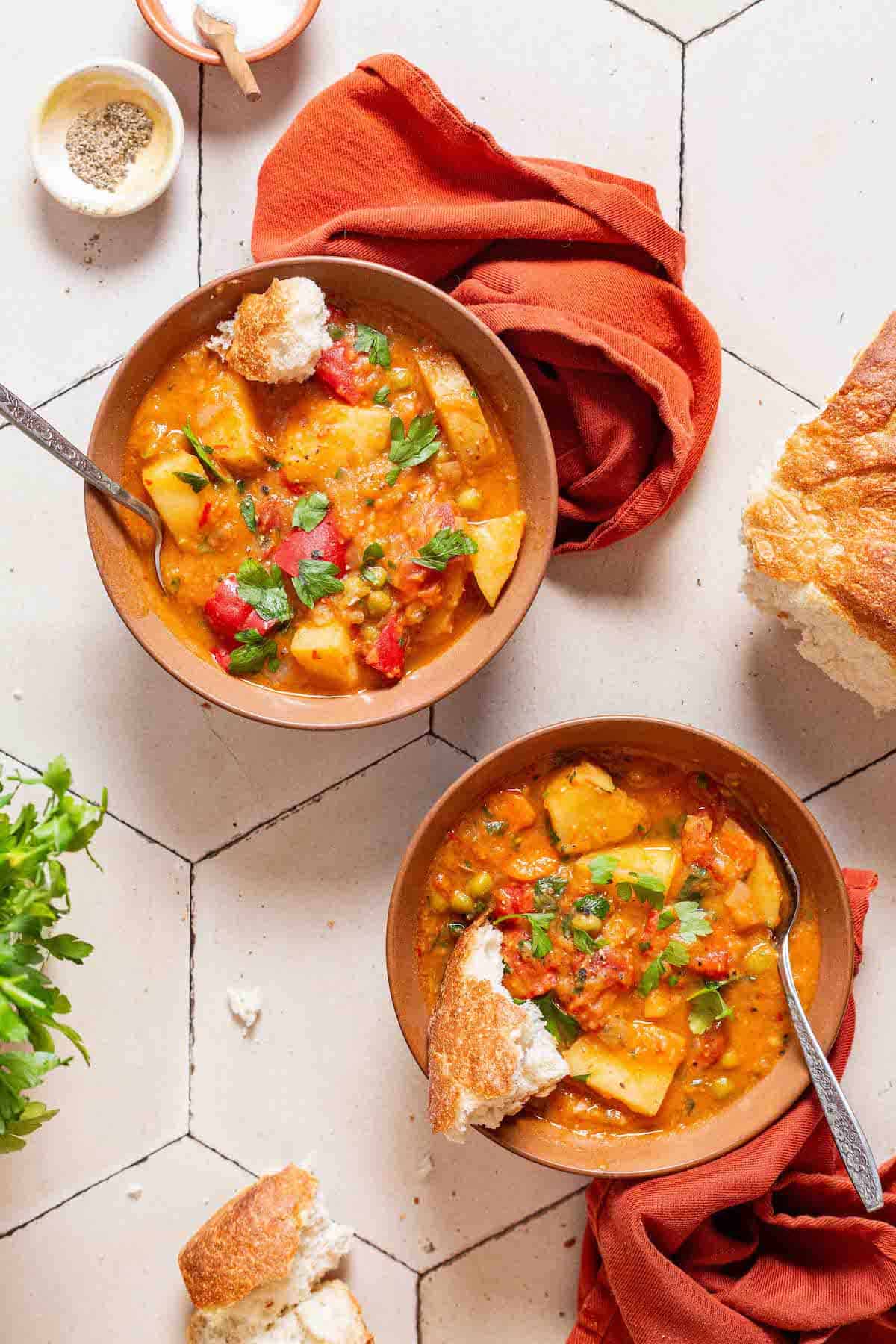 An overhead photo of two bowls of vegetable stew with spoons next to small bowls of salt and pepper, parsley and some crusty bread.