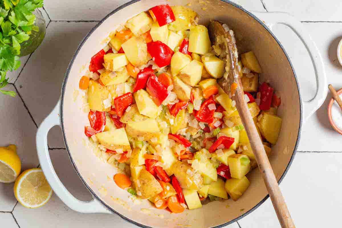 An overhead photo of onions, garlic, celery, carrots, potatoes and a bell pepper being sauteed in a large pot with a wooden spoon. There is a small bowl of salt, two lemon halves and parsley surrounding the pot.