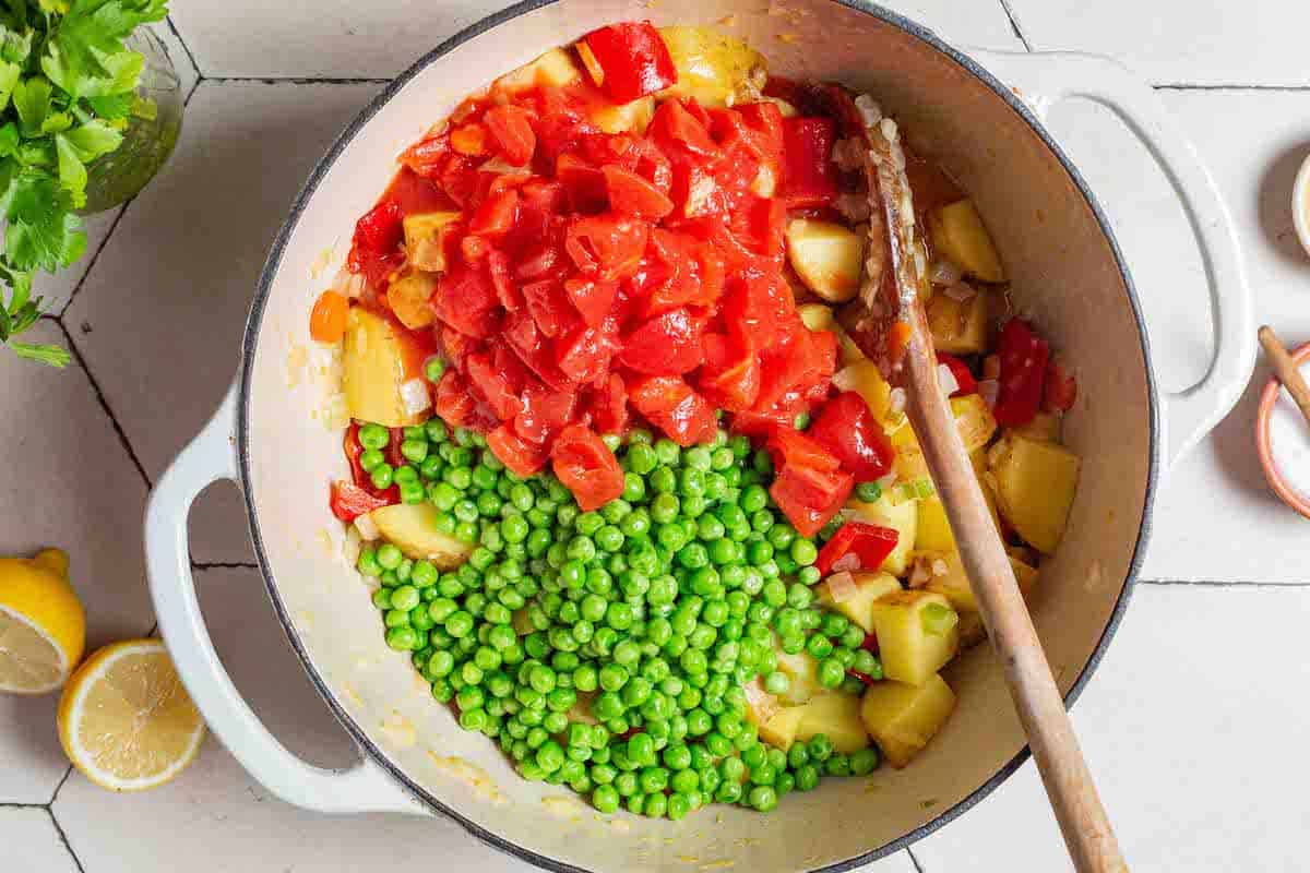An overhead photo of the peas and diced tomatoes added to the other ingredients in the large pot with a wooden spoon. There is a small bowl of salt, two lemon halves and parsley surrounding the pot.