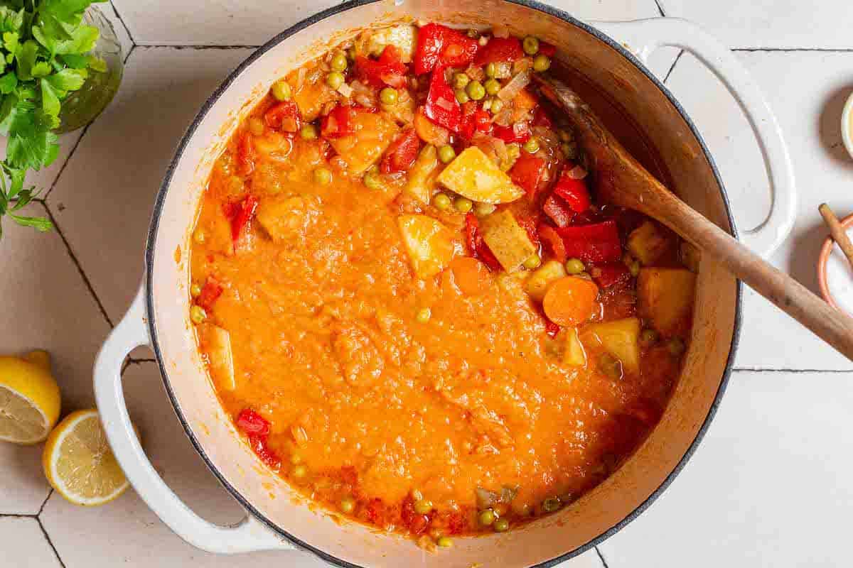 An overhead photo of the vegetable stew in a large pot after some of it has been mixed with an immersion blender. There is a small bowl of salt, 2 lemon halves and parsley surrounding the pot.