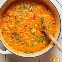 An overhead photo of vegetable stew in a large pot with a wooden spoon next to small bowls of salt and pepper, 2 lemon halves and parsley.