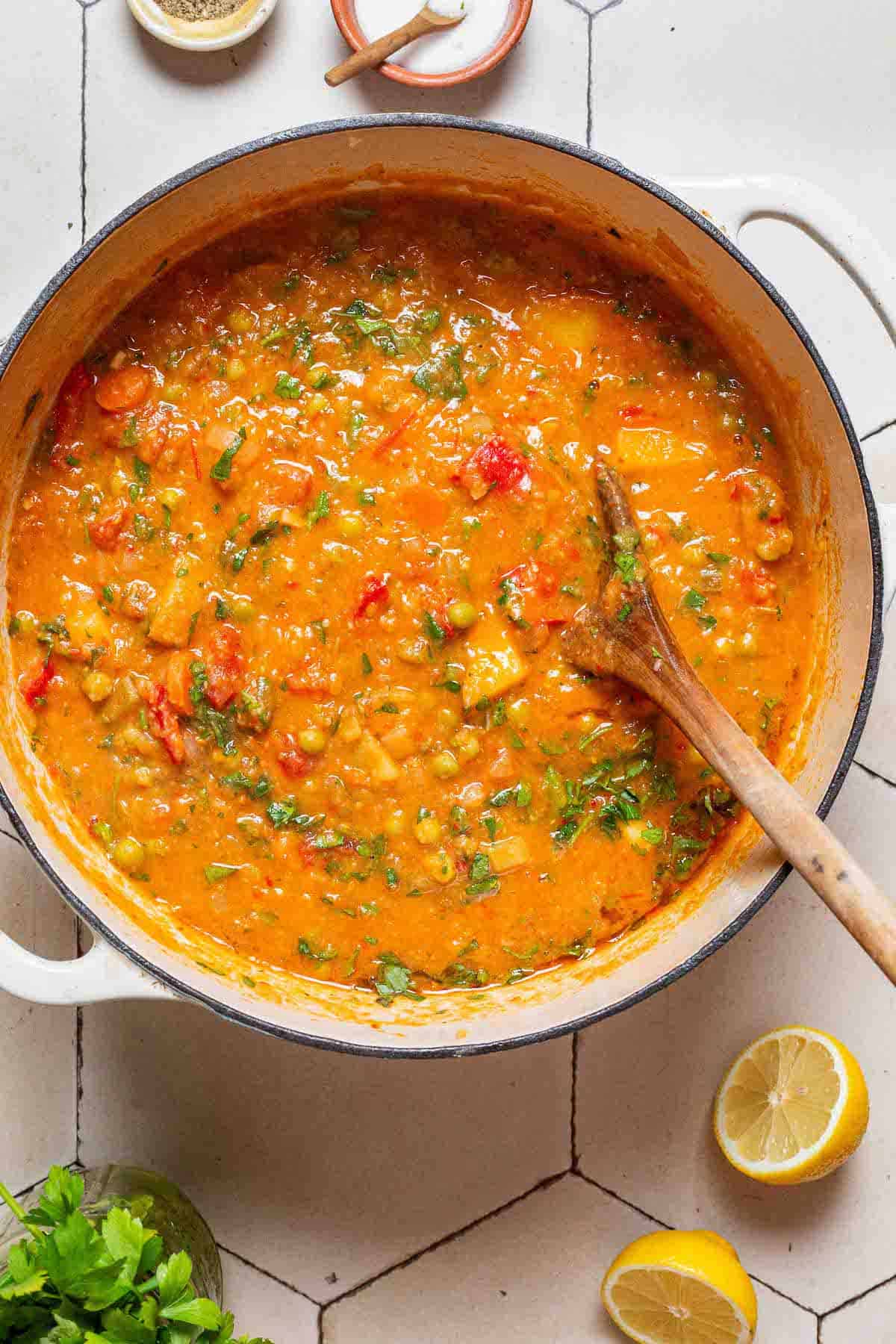 An overhead photo of vegetable stew in a large pot with a wooden spoon next to small bowls of salt and pepper, 2 lemon halves and parsley.