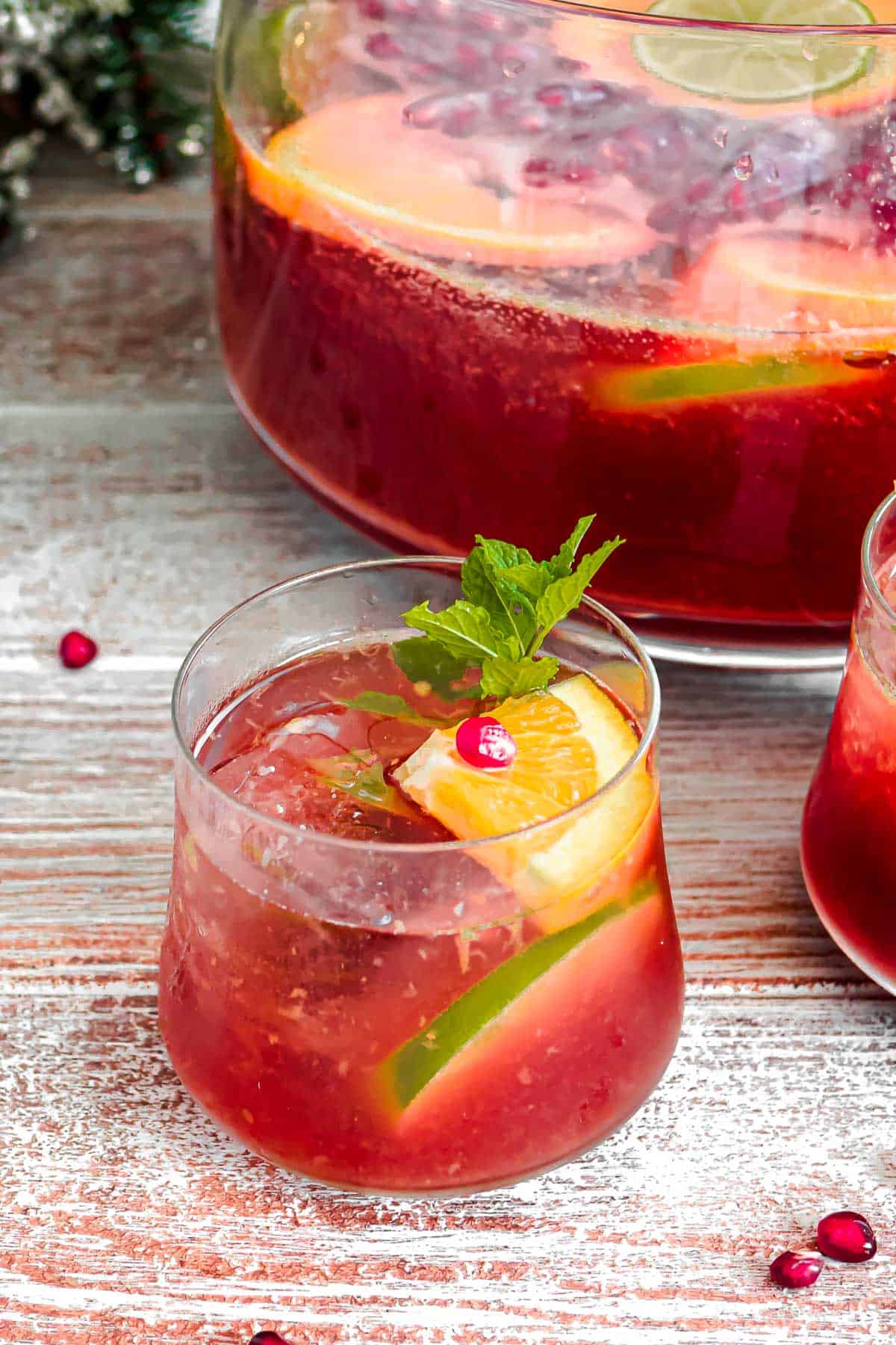 a glass of holiday punch garnished with orange and lime slices and mint, in front of a bowl of the punch.