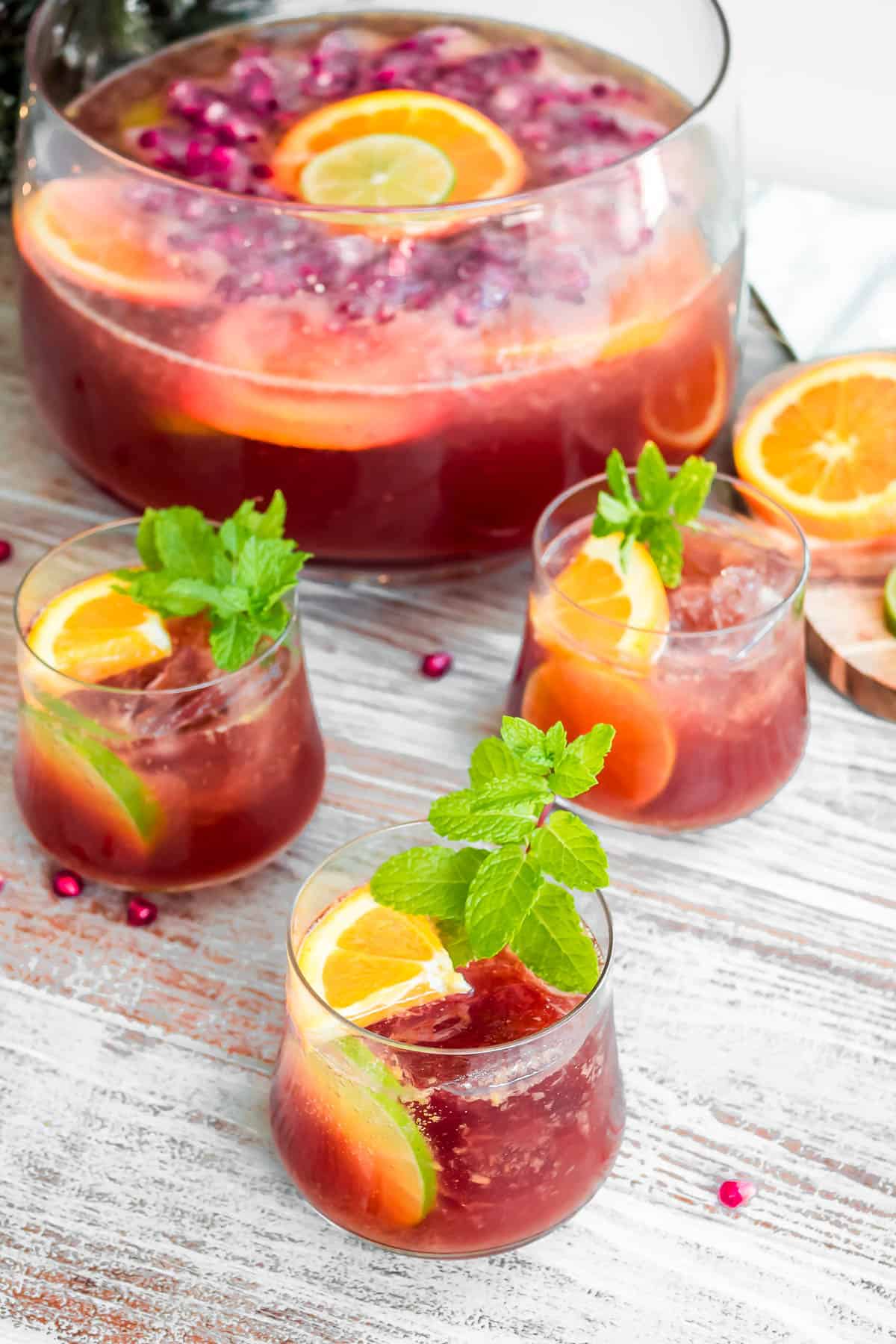 three glasses of holiday punch garnished with orange and lime slices and mint, in front of a bowl of the punch, and a cutting board containing more slices oranges.