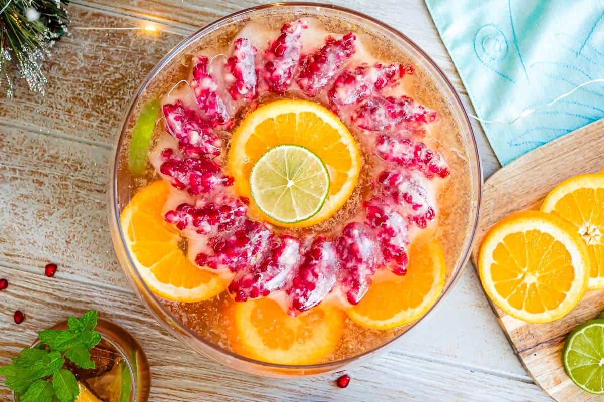 an overhead photo of holiday punch in a bowl with a pomegranate ice ring and garnishes of slices of oranges of limes next to a glass of holiday punch and a cutting board with more orange and lime slices.