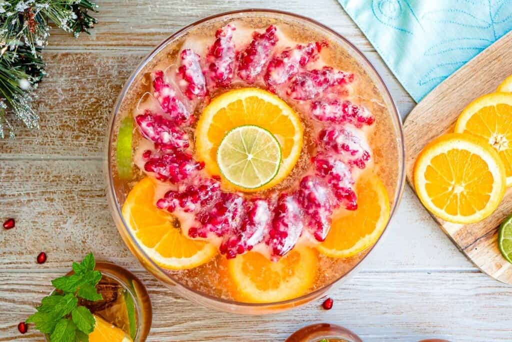 an overhead photo of holiday punch in a bowl with a pomegranate ice ring and garnishes of slices of oranges of limes next to a glass of holiday punch and a cutting board with more orange and lime slices.
