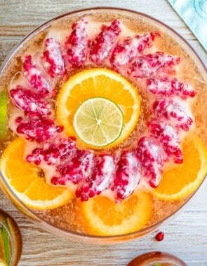 an overhead photo of holiday punch in a bowl with a pomegranate ice ring and garnishes of slices of oranges of limes next to a glass of holiday punch and a cutting board with more orange and lime slices.