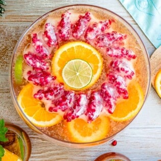 an overhead photo of holiday punch in a bowl with a pomegranate ice ring and garnishes of slices of oranges of limes next to a glass of holiday punch and a cutting board with more orange and lime slices.
