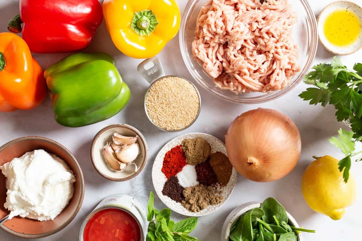 ingredients for chicken stuffed peppers including bell peppers, bulgur wheat, olive oil, onion, garlic, ground chicken, salt, aleppo pepper, coriander, cumin, sweet paprika, sumac, nutmeg, diced tomatoes, baby spinach, parsley, mint, greek yogurt, and lemon.